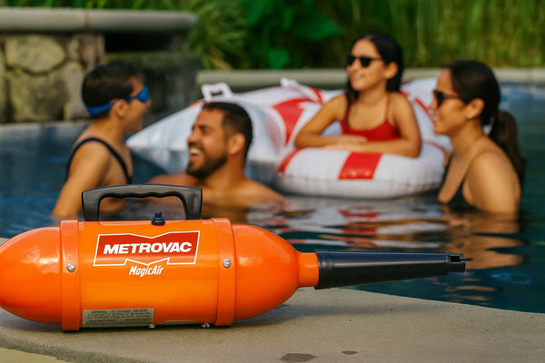 An orange Metrovac MagicAir inflator/deflator sits on the edge of a swimming pool. In the background, three people are smiling and relaxing in the water, with one person in an inflatable pool float.