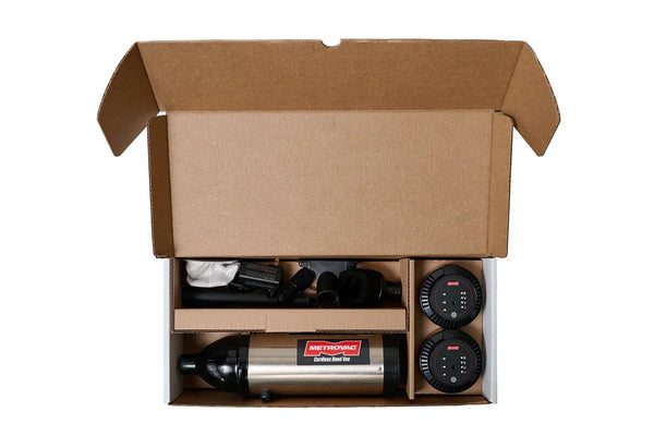Open cardboard box showing MetroVac Cordless Hand Vac, two rechargeable batteries, and multiple attachments neatly packed for shipping.