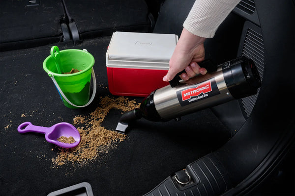 Person using MetroVac Cordless Hand Vac to clean sand from car trunk after a beach trip, next to a cooler and toy bucket.