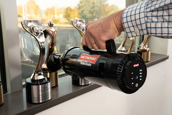 Hand holding the MetroVac Cordless DataVac CDV-1-A to clean dust from award trophies displayed on a shelf.