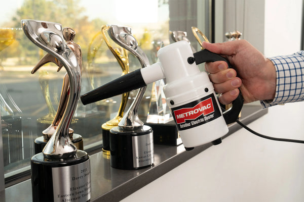 A person uses the MetroVac DATAVAC® ELECTRIC DUSTER® to clean a display of metallic trophies, showcasing the device's utility for gently dusting delicate or intricate surfaces.