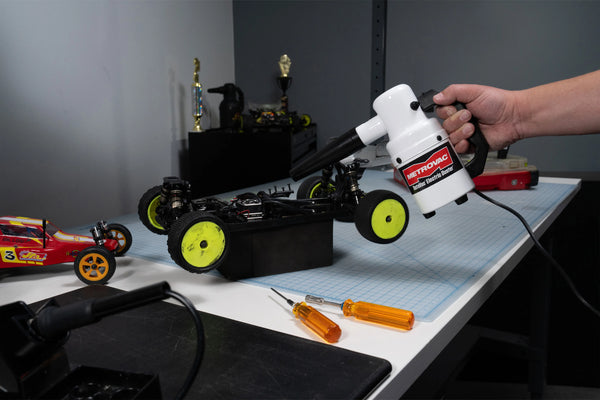 A person uses the MetroVac DATAVAC® ELECTRIC DUSTER® to clean a dust-covered RC car on a workbench, surrounded by tools and model cars in a hobby workspace.